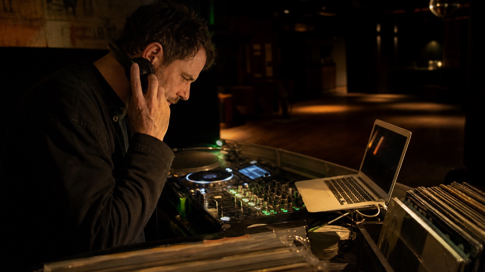 An experienced house DJ listens closely while checking a playlist beside professional decks